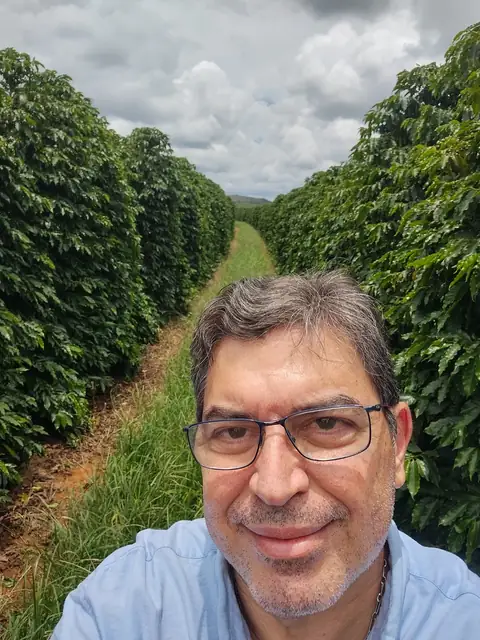 Fernando Reis de óculos e camisa azul-clara, numa selfie entre duas fileiras de cafeeiros, com o céu nublado ao fundo